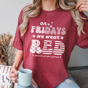 Peut inclure: Un t-shirt rouge avec un motif blanc qui dit "On Fridays We Wear Red" avec des &eacute;toiles et des rayures. Le texte "Remember Everyone Deployed" est sous le motif.