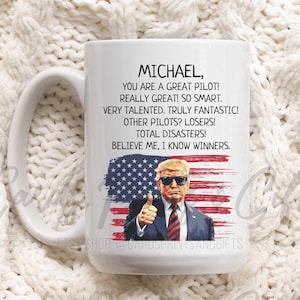 May include: White ceramic mug with a handle. The mug has a graphic of a man wearing a suit and sunglasses in front of an American flag. The text on the mug reads "MICHAEL, YOU ARE A GREAT PILOT! REALLY GREAT! SO SMART. VERY TALENTED. TRULY FANTASTIC! OTHER PILOTS? LOSERS! TOTAL DISASTERS! BELIEVE ME, I KNOW WINNERS."