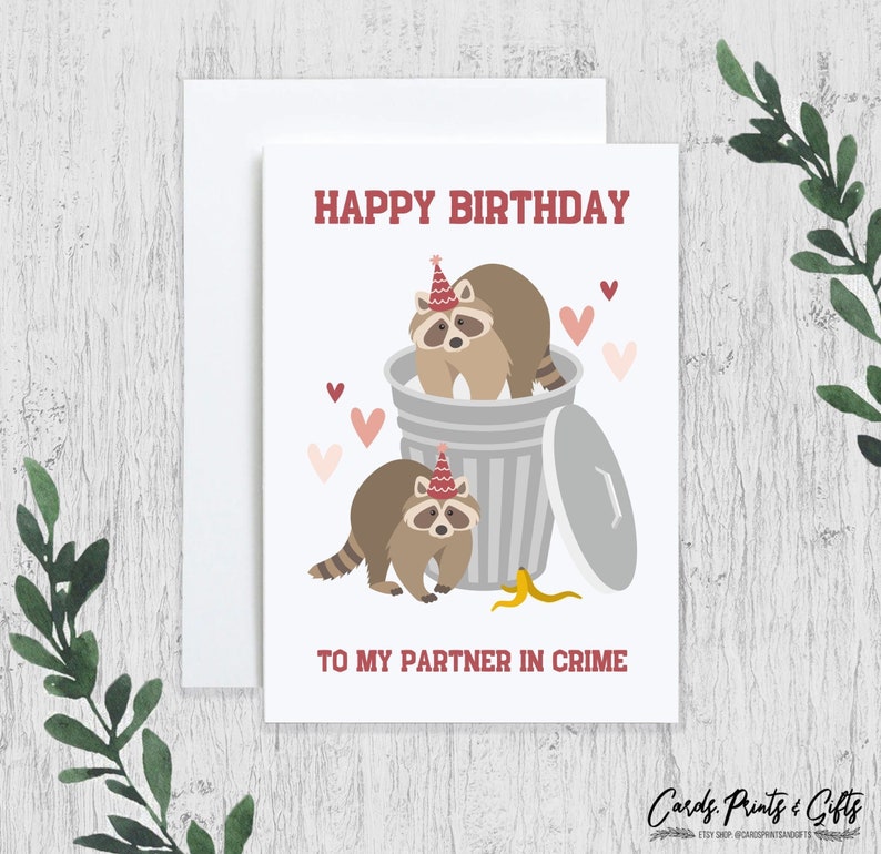 May include: Happy birthday to my partner in crime card featuring two raccoons wearing party hats and a bin with a banana peel. The card has a red and white colour scheme with pink hearts.