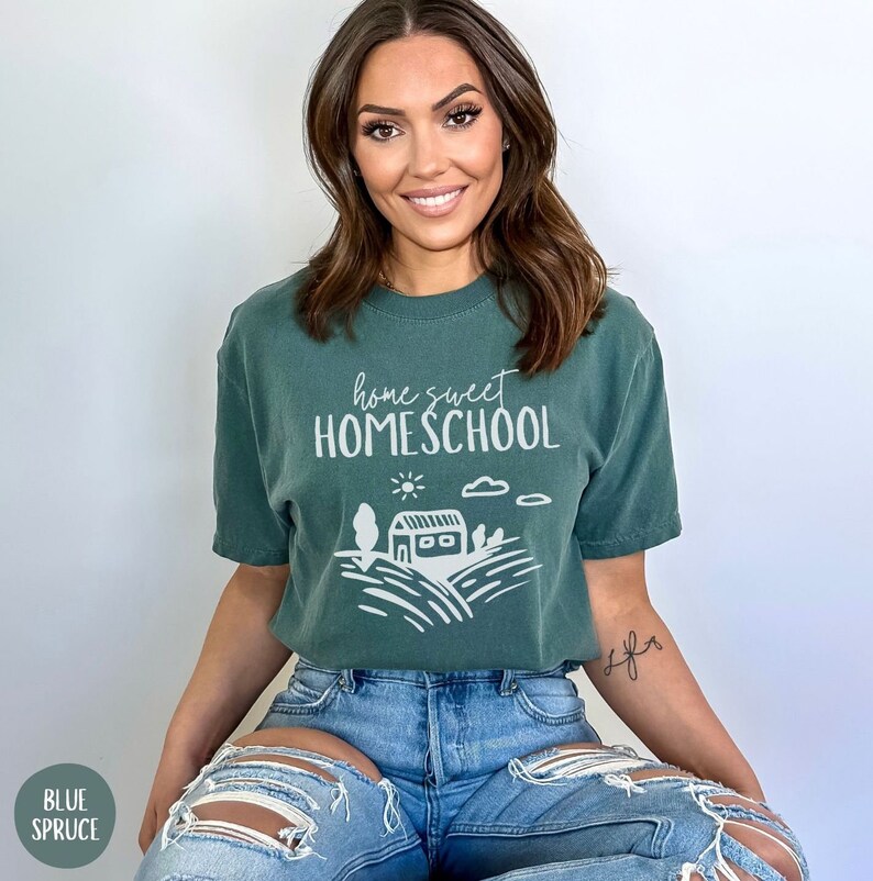 May include: A woman wearing a green t-shirt with the text "home sweet homeschool" and a white graphic of a house, sun, and clouds. She is wearing blue jeans with rips in the knees.