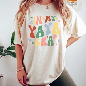Puede incluir: Una mujer lleva una camiseta de color crema con el texto "IN MY YAYA ERA" en letras de colores de estilo retro. La camiseta tiene un corte holgado y est&aacute; decorada con peque&ntilde;os gr&aacute;ficos de flores y estrellas.