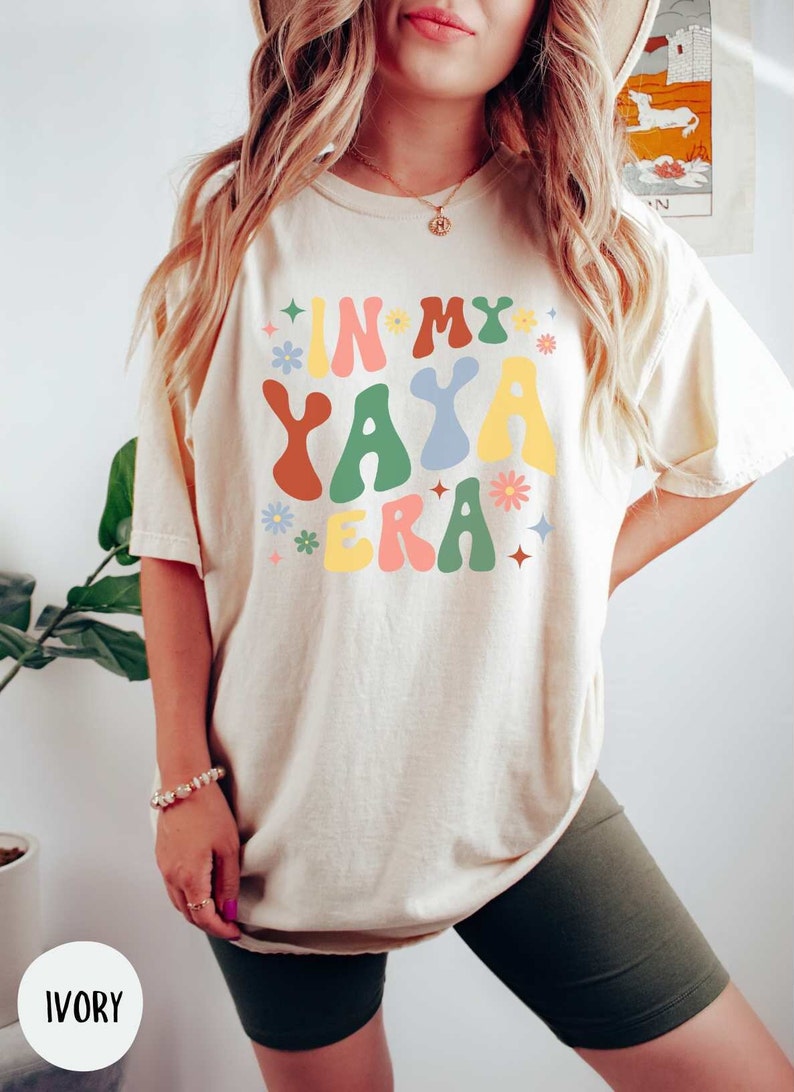 Puede incluir: Una mujer lleva una camiseta de color crema con el texto "IN MY YAYA ERA" en letras de colores de estilo retro. La camiseta tiene un corte holgado y est&aacute; decorada con peque&ntilde;os gr&aacute;ficos de flores y estrellas.