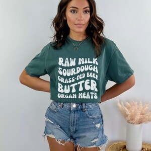 May include: A woman wearing a teal green t-shirt with white text that reads "RAW MILK SOURDOUGH GRASS-FED BEEF BUTTER ORGAN MEATS". She is also wearing denim shorts.