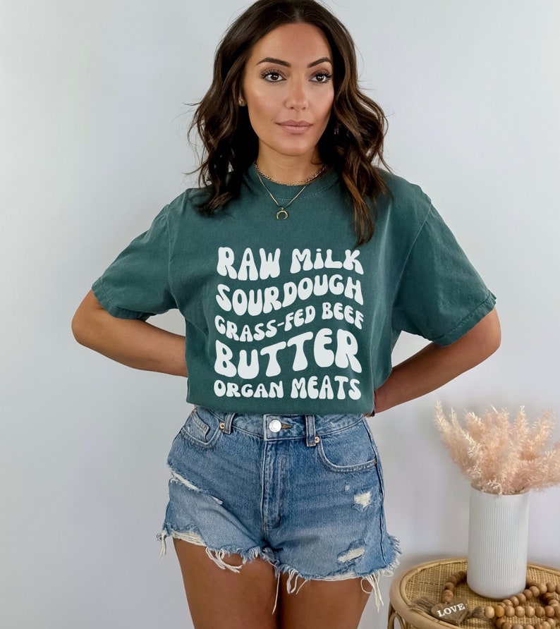 May include: A woman wearing a teal green t-shirt with white text that reads "RAW MILK SOURDOUGH GRASS-FED BEEF BUTTER ORGAN MEATS". She is also wearing denim shorts.