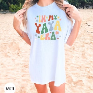 Puede incluir: Camiseta blanca con el texto "In My Yaya Era" en letras onduladas de colores con flores y estrellas alrededor del texto.