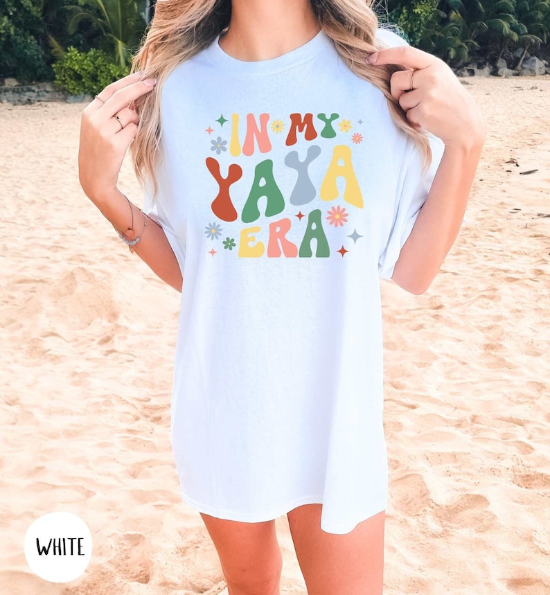 Puede incluir: Camiseta blanca con el texto "In My Yaya Era" en letras onduladas de colores con flores y estrellas alrededor del texto.