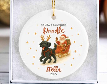 May include: White ceramic Christmas ornament featuring a black poodle wearing reindeer antlers pulling Santa's sleigh. The ornament has the text "SANTA'S FAVORITE DOODLE" and the name "Stella" with the year "2025".
