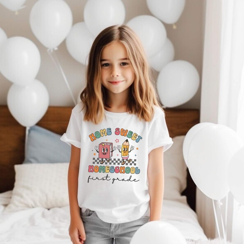 May include: A white t-shirt with a colorful graphic that says "Home Sweet Homeschool First Grade". The graphic features a cartoon book and pencil.
