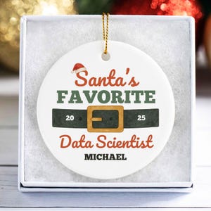 May include: A white ceramic Christmas ornament with a gold string, presented in a white box. The ornament features the text "Santa's FAVORITE Data Scientist" with a Santa hat, belt design, and the name "MICHAEL".