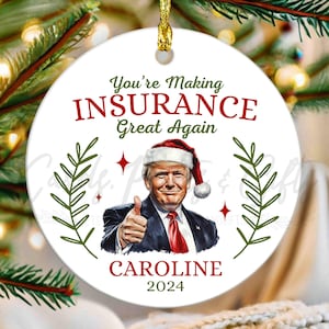 May include: White round Christmas ornament with a red and white Santa hat wearing Donald Trump caricature. The ornament has the text "You're Making Insurance Great Again" and the name "CAROLINE 2024".