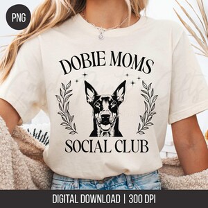 Puede incluir: Camiseta color crema con el texto "DOBIE MOMS SOCIAL CLUB" en negro. Un dibujo en blanco y negro de un Doberman Pinscher está centrado en una corona de laurel. En la parte inferior se lee "DIGITAL DOWNLOAD | 300 DPI".