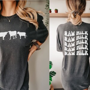 May include: A dark gray t-shirt with a white graphic of three cows on the front and the text "RAW MILK" repeated five times on the back.