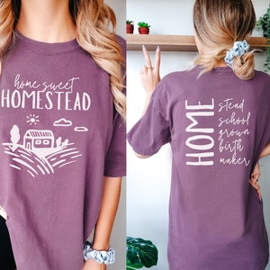 May include: A purple t-shirt with the text "home sweet homestead" and a white illustration of a house and fields on the front. The back of the shirt has the text "HOME" vertically stacked with the words "stead", "school", "grown", "birth", and "maker" stacked below it.