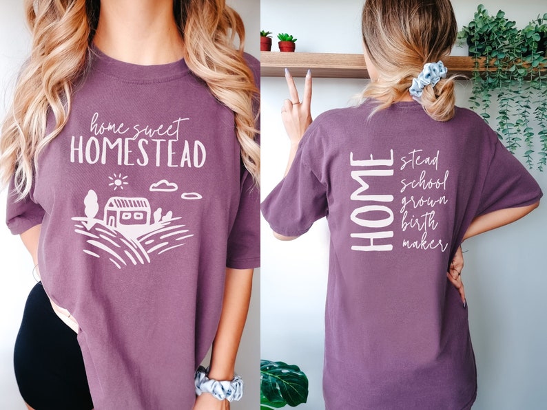 May include: A purple t-shirt with the text "home sweet homestead" and a white illustration of a house and fields on the front. The back of the shirt has the text "HOME" vertically stacked with the words "stead", "school", "grown", "birth", and "maker" stacked below it.