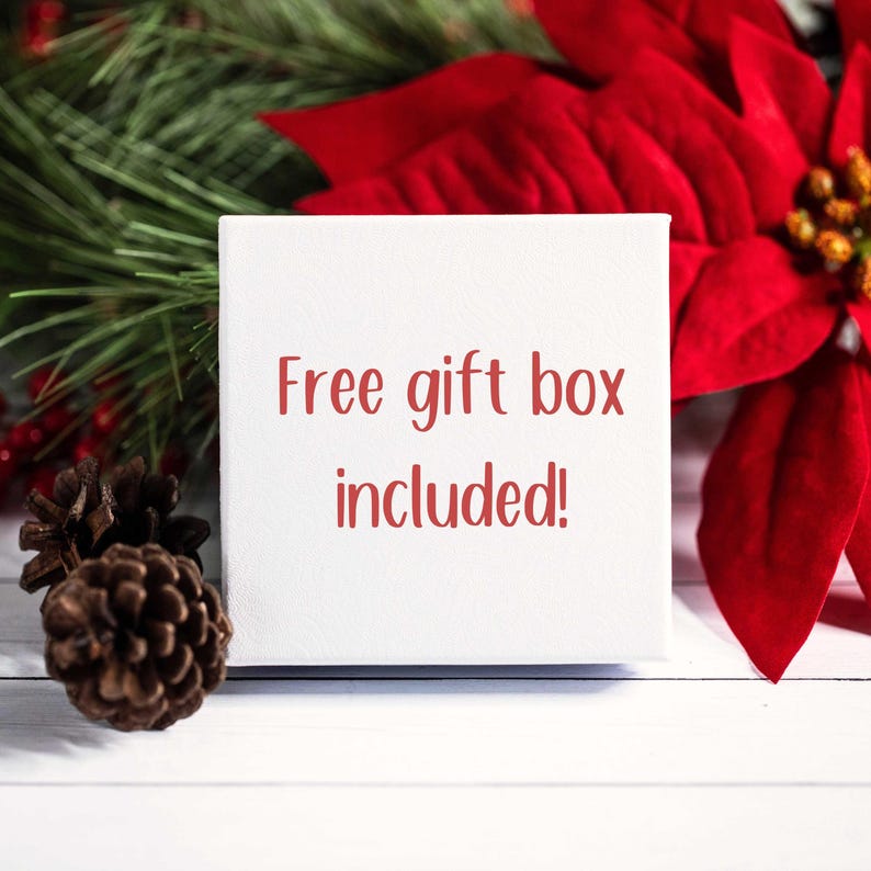 Op de afbeelding: Een witte geschenkdoos met de rode tekst "Free gift box included!". De doos wordt omringd door dennennaalden, een dennenappel en een rode poinsettia bloem, wat een feestelijke sfeer oproept.