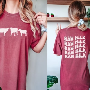 May include: A maroon t-shirt with a white silhouette of three cows on the front and the text "RAW MILK" repeated five times on the back.
