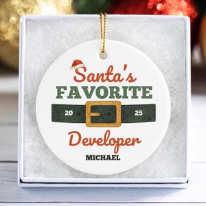 May include: A white ceramic Christmas ornament with the text "Santa's Favorite Developer" in red and green. The ornament features a black belt design with a gold buckle and the name "MICHAEL" at the bottom.