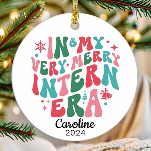 May include: White round Christmas ornament with the text "In My Very Merry Intern Era" in colorful retro font. The ornament also includes a small Christmas tree, snowflakes, and bells. The name "Caroline" and the year "2024" are written below the text.
