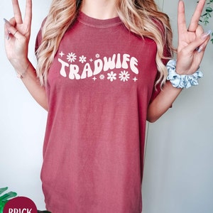 Puede incluir: Una camiseta de gran tama&ntilde;o de color rojo ladrillo con el texto "TRADWIFE" en blanco con una fuente retro y detalles de flores blancas.