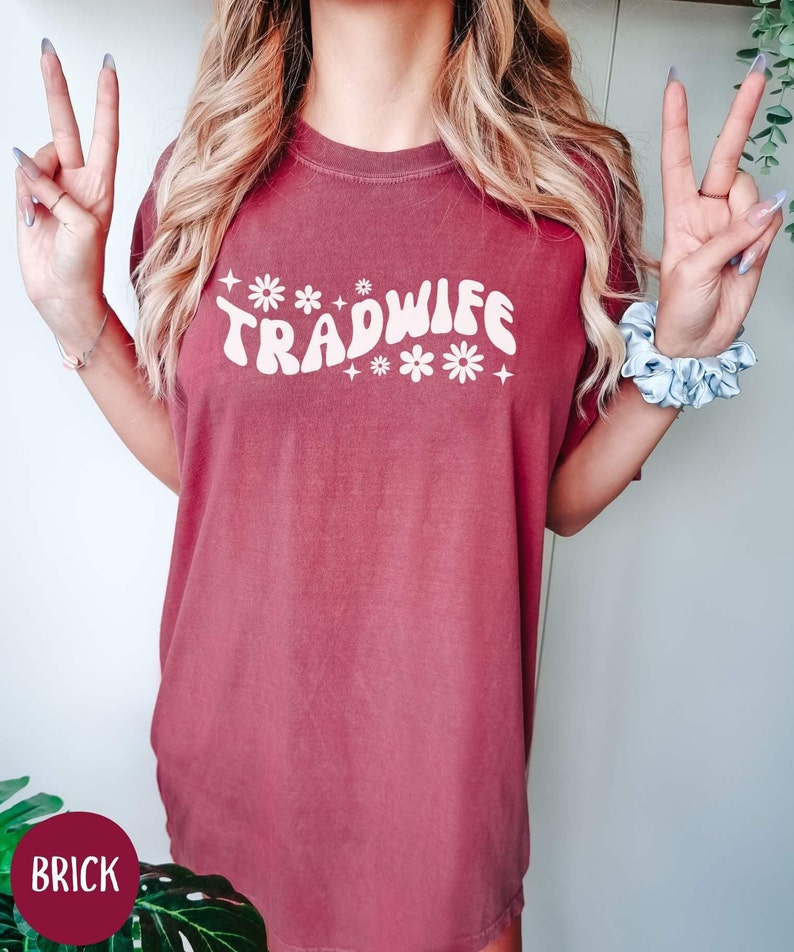 Puede incluir: Una camiseta de gran tama&ntilde;o de color rojo ladrillo con el texto "TRADWIFE" en blanco con una fuente retro y detalles de flores blancas.