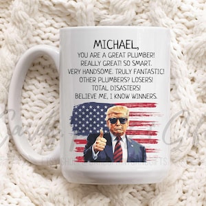 May include: White ceramic coffee mug with a red, white, and blue American flag design. The mug has a handle and features a text overlay that reads "MICHAEL, YOU ARE A GREAT PLUMBER! REALLY GREAT! SO SMART. VERY HANDSOME. TRULY FANTASTIC! OTHER PLUMBERS? LOSERS! TOTAL DISASTERS! BELIEVE ME, I KNOW WINNERS."