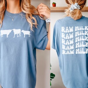 May include: A light blue t-shirt with a white graphic of three cows on the front and the text "RAW MILK" repeated five times on the back.