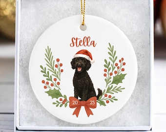 May include: A white ceramic Christmas ornament featuring a black dog wearing a Santa hat, the name "Stella", and the year "2025". The ornament is decorated with a wreath of green leaves and red berries, and a red bow.