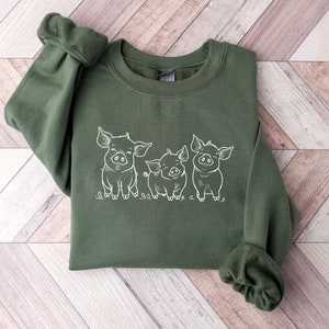 May include: A green sweatshirt with a white outline of three pigs on the front. The pigs are facing forward and have their snouts pointed towards the viewer. The sweatshirt is folded in half and the sleeves are rolled up.