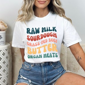 May include: White t-shirt with a colorful graphic that says "Raw Milk Sourdough Grass-Fed Beef Butter Organ Meats".