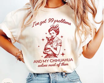 May include: A white t-shirt with a red graphic print of a woman holding a chihuahua. The text on the shirt reads "I've got 99 problems and my chihuahua solves most of them".