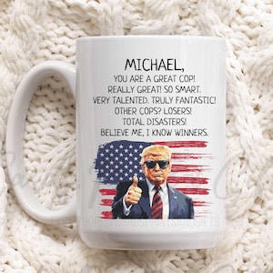 May include: White ceramic coffee mug with a black and red American flag design. The mug has a handle and text that reads "MICHAEL, YOU ARE A GREAT COP! REALLY GREAT! SO SMART. VERY TALENTED. TRULY FANTASTIC! OTHER COPS? LOSERS! TOTAL DISASTERS! BELIEVE ME, I KNOW WINNERS."