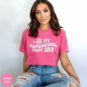 May include: A woman wearing a pink t-shirt with the text "In My Homeschool Mom Era" in white. She is sitting with her legs crossed and wearing blue jeans.