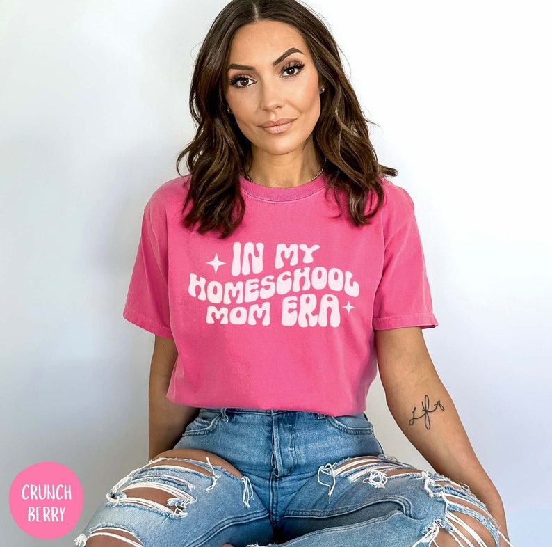 May include: A woman wearing a pink t-shirt with the text "In My Homeschool Mom Era" in white. She is sitting with her legs crossed and wearing blue jeans.