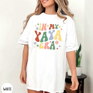 Puede incluir: Camiseta blanca de gran tama&ntilde;o con el texto "IN MY YAYA ERA" en letras de colores de estilo retro con flores y estrellas.