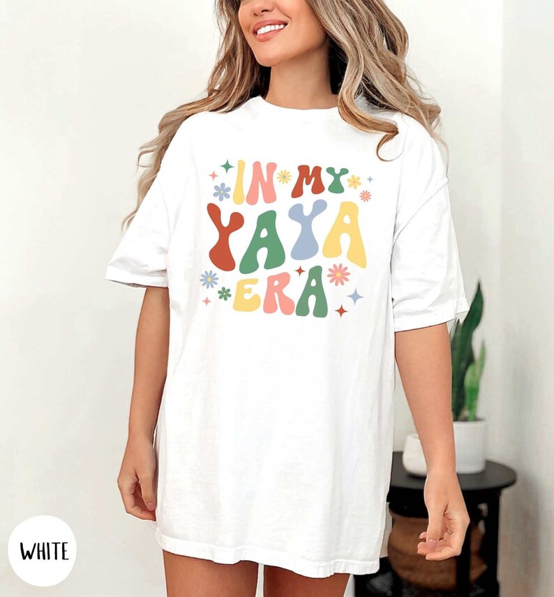 Puede incluir: Camiseta blanca de gran tama&ntilde;o con el texto "IN MY YAYA ERA" en letras de colores de estilo retro con flores y estrellas.