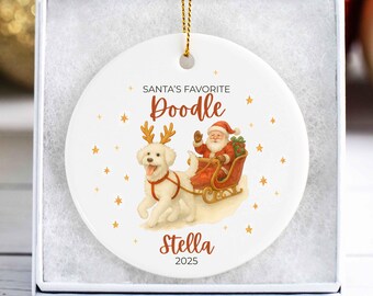 May include: White ceramic Christmas ornament with a gold string, featuring a Doodle dog wearing antlers pulling Santa's sleigh. The text reads "Santa's Favorite Doodle" and "Stella 2025". The ornament is in a white gift box.