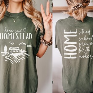 May include: A green t-shirt with the text "home sweet homestead" on the front and a stylized illustration of a house and fields. The back of the shirt has the word "HOME" vertically down the left side and the words "stead, school, grown, birth, maker" vertically down the right side.