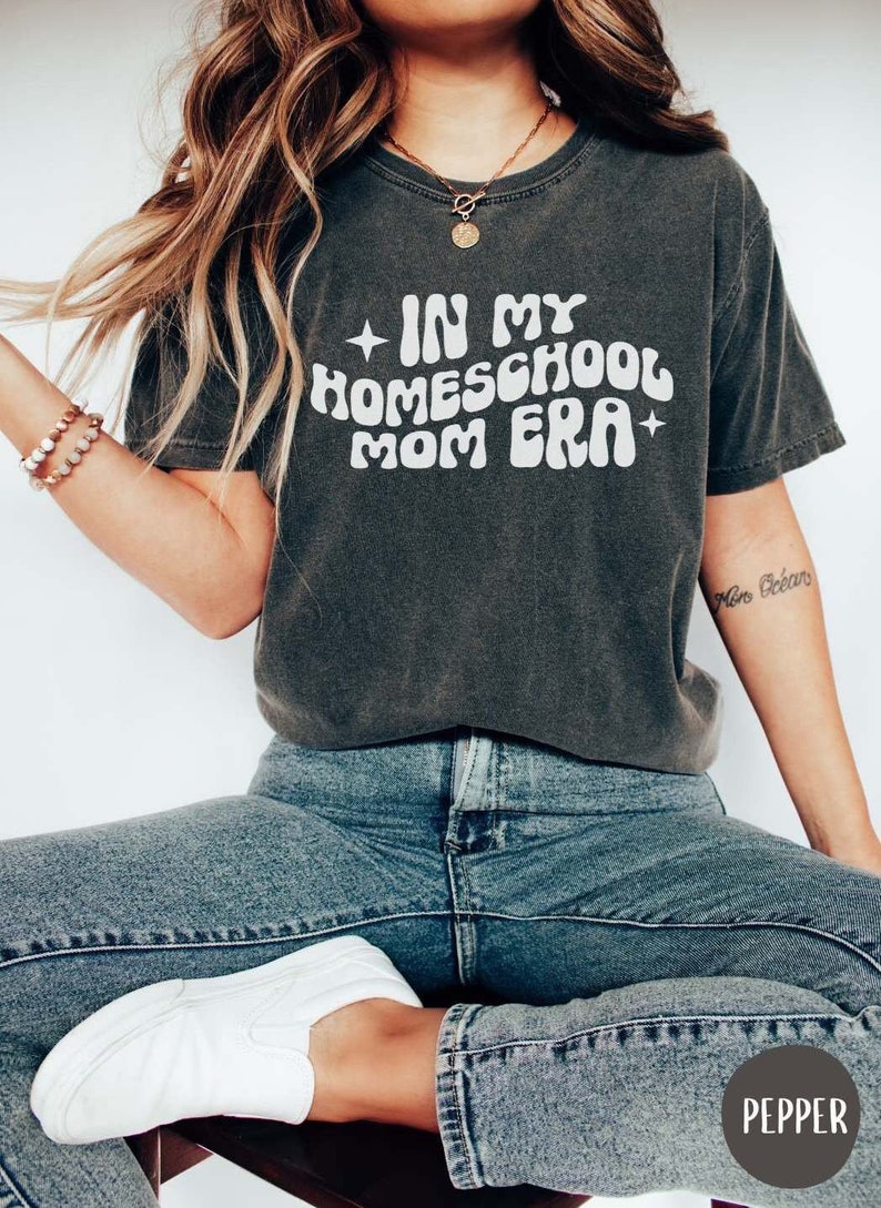 May include: A dark gray t-shirt with the text "In My Homeschool Mom Era" in white, retro-style lettering. The shirt is worn by a person with long, wavy hair and a necklace.