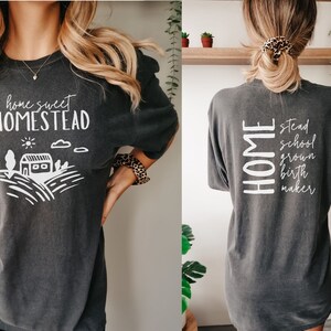 May include: A dark gray t-shirt with a white graphic on the front that says "home sweet homestead" and a small illustration of a house and fields. The back of the shirt says "HOME stead school grown birth maker".