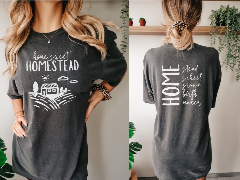 May include: A dark gray t-shirt with a white graphic on the front that says "home sweet homestead" and a small illustration of a house and fields. The back of the shirt says "HOME stead school grown birth maker".