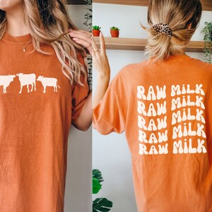 May include: Orange t-shirt with a white silhouette of three cows on the front and the text "RAW MILK" repeated five times in a retro font on the back.
