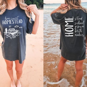 May include: A dark gray t-shirt with a white graphic on the front that says "home sweet homestead" and a white graphic on the back that says "HOME" with the words "stead", "school", "grown", "birth", and "maker" stacked vertically below it.