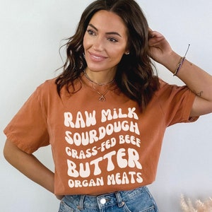 May include: A woman wearing a burnt orange t-shirt with the text "RAW MILK SOURDOUGH GRASS-FED BEEF BUTTER ORGAN MEATS" in white.