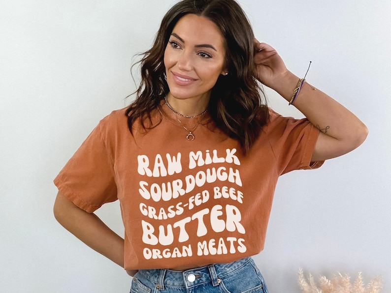 May include: A woman wearing a burnt orange t-shirt with the text "RAW MILK SOURDOUGH GRASS-FED BEEF BUTTER ORGAN MEATS" in white.