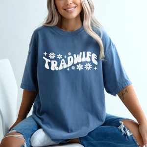 Puede incluir: Una camiseta azul con un gr&aacute;fico blanco que dice "TRADWIFE" en una fuente retro con flores y estrellas.