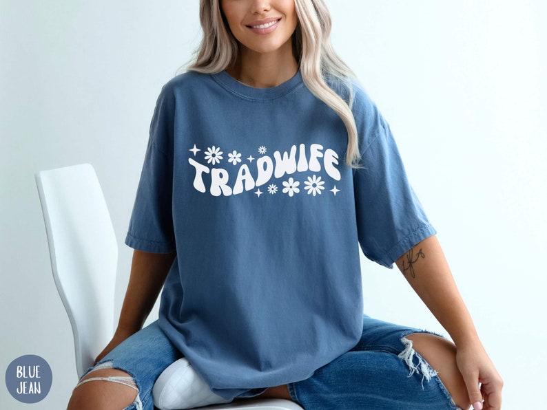 Puede incluir: Una camiseta azul con un gr&aacute;fico blanco que dice "TRADWIFE" en una fuente retro con flores y estrellas.
