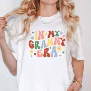May include: White t-shirt with the text "In My Granny Era" in colorful retro font with flowers and stars.