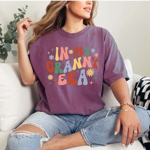 May include: A purple t-shirt with the text "In My Granny Era" in a retro font with colorful flowers.