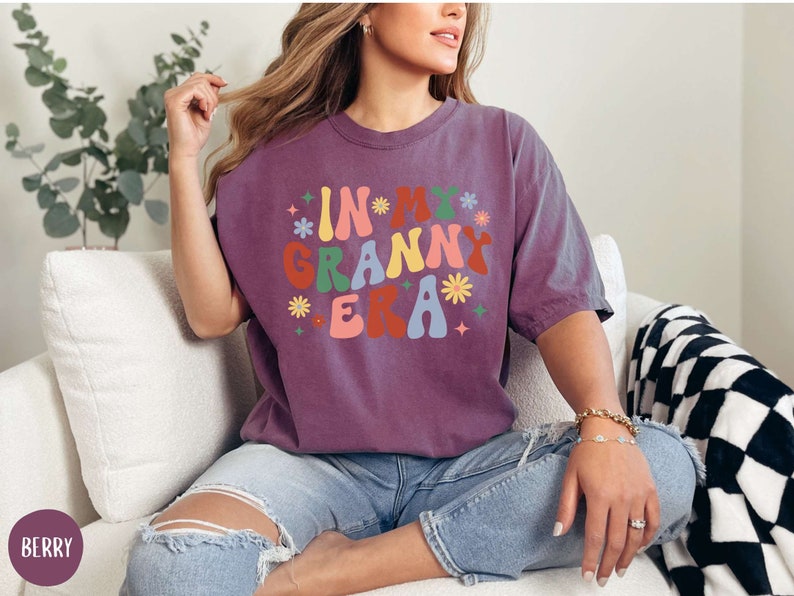 May include: A purple t-shirt with the text "In My Granny Era" in a retro font with colorful flowers.