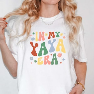 Puede incluir: Camiseta blanca con un gr&aacute;fico colorido que dice "In My Yaya Era" con flores y estrellas.